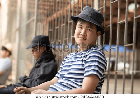 Asian teenboy in a black and white t-shir sits with his knees pressed against a metal fence panel in a juvenile detention facility, awaiting further release, freedom and detention of people concept. Royalty-Free Stock Photo #2434644863