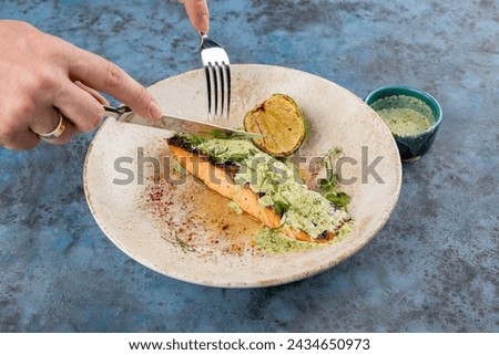 Woman's hand is using a knife and fork to cut Salmon steak grilled in a white plate on blue background