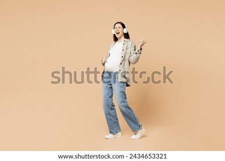 Full body young pregnant woman future mom wear grey shirt headphones with stomach tummy with baby listen music play air guitar isolated on plain beige background. Maternity family pregnancy concept