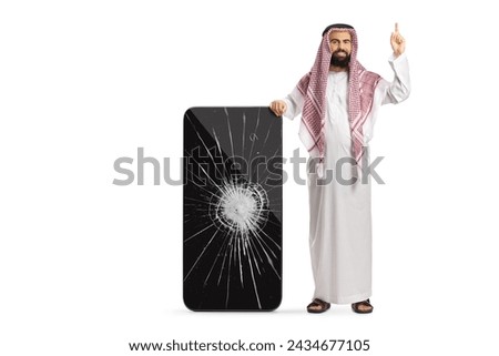 Saudi arab man in ethnic clothes leaning on a mobile phone with cracked screen and pointing up isolated on white background