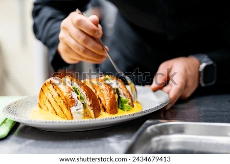 A close-up of a chef garnishing a grilled sandwich with fresh ingredients, drizzling sauce. The golden-brown bread reveals lettuce and other fillings. The background suggests a kitchen setting.
