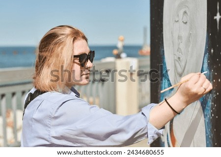 Young female painter in sunglasses passionately paints picture with paintbrush for outdoor street exhibition, female artist engrossed in creating vibrant artwork at bright sunny day