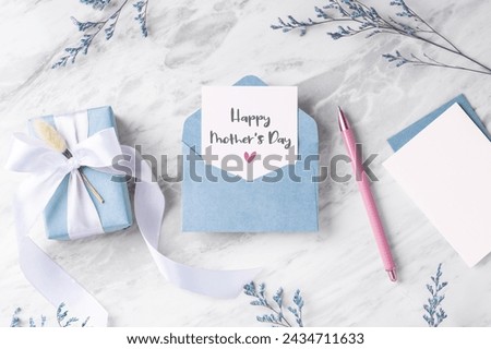 Mother's day card with blue gift box and envelope on marble table with flower and pen, top view