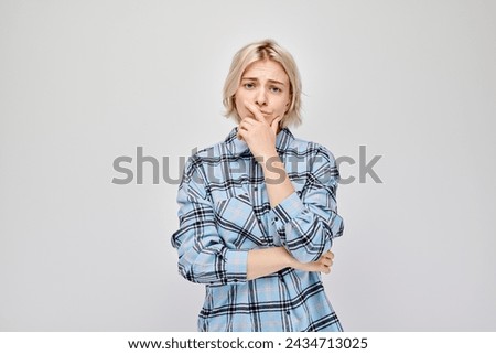 Young woman in plaid shirt looking uncertain, hand on chin, against a grey background.