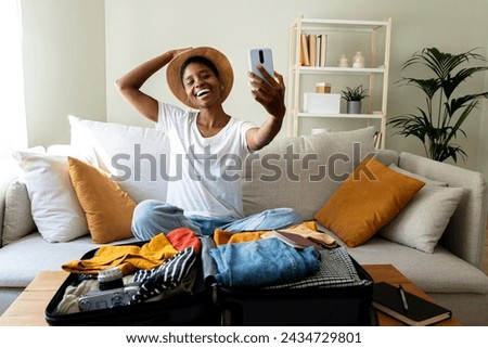 Happy young African American woman sitting on sofa packing suitcase for summer holidays abroad wearing hat taking selfie with mobile phone. Vacation concept.