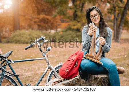 Young female student using mobile phone while studying for exam in public park during the sunny Autumn day.
