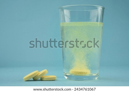 Photograph of orange colour water soluble vitamin C dissolving in glass of water with bubbles and pile of pills or tablets on blue colour background