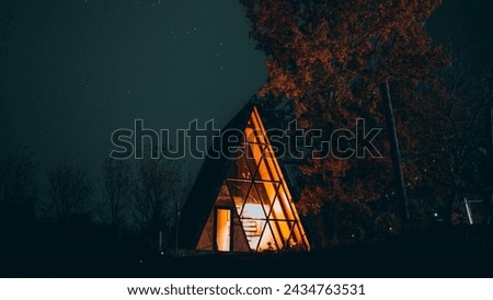 The image is of a house with a large triangular roof, situated outdoors at night under a starry sky. The landscape includes trees and a light illuminating the scene.