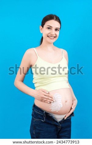 Close up of pregnant woman in opened jeans applying skin care cream on her belly to prevent stretch marks at colorful background with copy space. Healthy skin concept.
