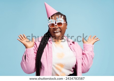 Beautiful excited African American woman wearing stylish party eyeglasses, birthday cone looking at camera. Stylish plus size female isolated on blue background. Concept of celebration, birthday