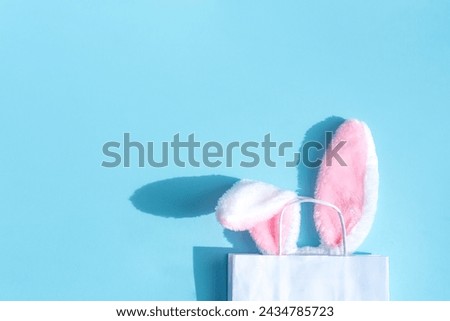 Fluffy bunny ears on pastel background sticking out of a white paper bag. Easter shopping concept. Top view, space for text.