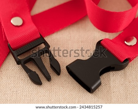 Two ends of a black contoured side release plastic buckle with red nylon belt on a beige textile background 