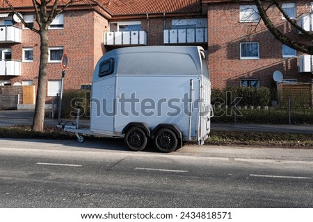 A small trailer for transporting a horse standing on the side of the road against the background of a brick building. Royalty-Free Stock Photo #2434818571
