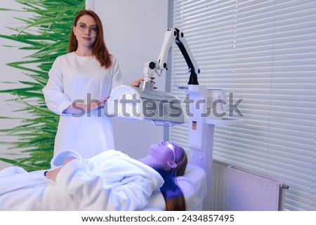 Portrait of beautician specialist connecting and adjusting innovation device for acne treatments and collagen production at aesthetic clinic. Woman lying during non-invasive type of phototherapy.