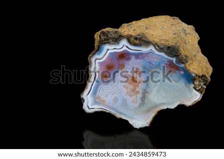 Polished cut of natural agate precious stone with raw brown surface on black background. Closeup of beautiful smooth icy blue cross section by chalcedony mineral rock with red stains and white border. Royalty-Free Stock Photo #2434859473