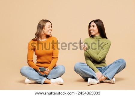 Full body young friends two women they wear orange green shirt casual clothes together sit hold in hand use show mobile cell phone isolated on plain pastel light beige background. Lifestyle concept