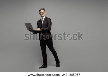 Full body side view adult employee IT business man corporate lawyer wears classic formal black suit shirt tie work in office hold use laptop pc computer look aside isolated on plain grey background Royalty-Free Stock Photo #2434865057
