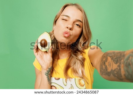 Close up young housewife housekeeper chef cook baker woman wear apron yellow t-shirt hold avocado do selfie shot mobile cell phone wink isolated on plain pastel green background. Cooking food concept