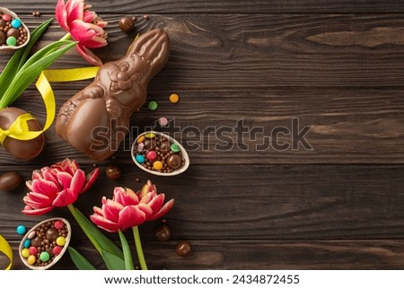 Joyful Easter confectionery display. Top view photo showing cracked open chocolate eggs, overflowing with rainbow candies, chocolate bunny, and lively tulips on timber table, blank area for messaging