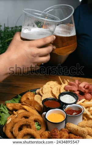 couple of a woman and a man in dark clothes eating on a date inside a Mexican wings and snacks bar, with wooden table and low lighting, they have beer in plastic cups and smile. Royalty-Free Stock Photo #2434895547