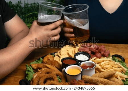 couple of a woman and a man in dark clothes eating on a date inside a Mexican wings and snacks bar, with wooden table and low lighting, they have beer in plastic cups and smile. Royalty-Free Stock Photo #2434895555