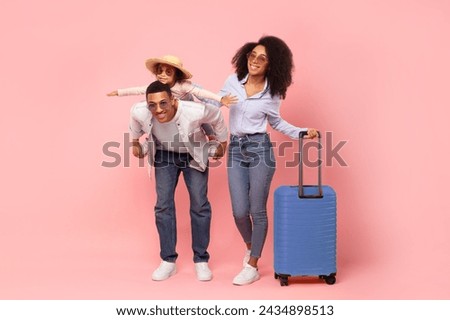 Excited black parents travelling with little daughter, dad piggybacking child girl, posing with suitcases on pink background in studio