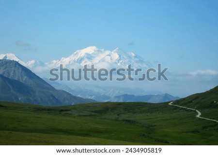 Magnificent vista of Denali framed by hills on a clear summer day with green vegetation foreground, a winding road in the distance, blue sky, and a snowy mountain summit rising above a bank of clouds. Royalty-Free Stock Photo #2434905819