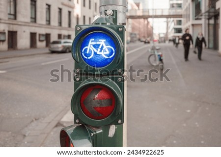 A close-up view of a traffic signal post at an urban intersection, showing a blue bicycle lane light illuminated alongside a red arrow indicating 'no left turn' for vehicles. 
