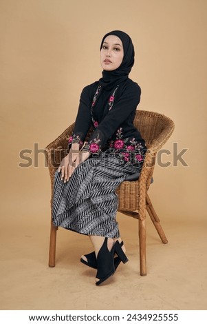 Beautiful Asian model wearing black kebaya dress with hijab, sitting happily on a rattan chair isolated over beige background. Eidul fitri festival, fashion and beauty concept.