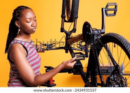 Youthful african american female carefully inspects the pedals and gear to make sure the bike is ready for outdoor activities. Sporty african american woman holding and checking foot brake of bicycle. Royalty-Free Stock Photo #2434931537
