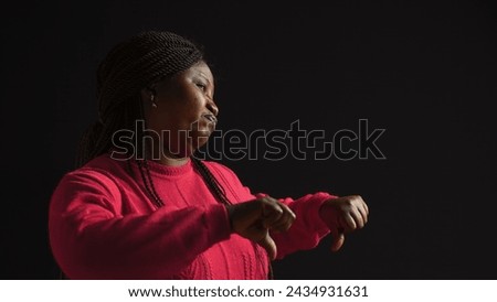 Standing against black background fashion model shows disapproval by holding her hands in front of her. African American woman in pink sweater uses thumbs down motion to disagree. Side-view shot. Royalty-Free Stock Photo #2434931631