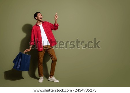 Full length photo of cool excited man dressed red shirt holding shoppers looking showing empty space isolated green color background