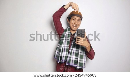 Excited Asian Muslim man holding and pointing at smartphone on white background