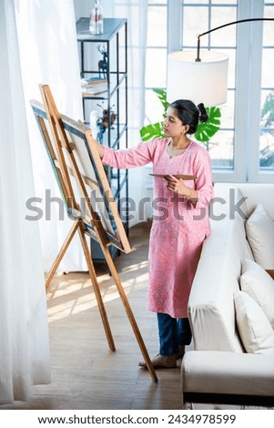 Cheerful Indian young artist girl painting picture at easel in living room