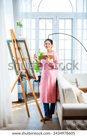Cheerful Indian young artist girl painting picture at easel in living room
