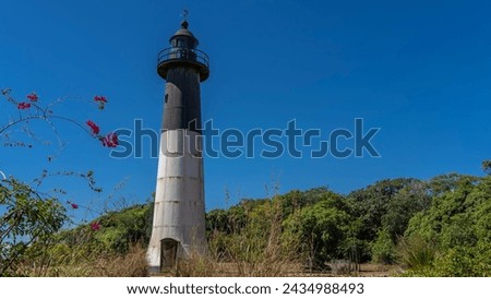 A tall old lighthouse on a tropical island. A black and white tower against a clear blue sky. Green vegetation all around. Madagascar. Nosy Iranja 