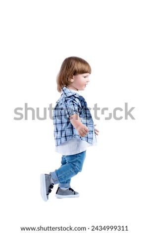 Smiling little boy 2 years old runs. Child wearing a blue checkered shirt and a white T-shirt. Activity, energy and childlike spontaneity. Full height. Side view. Royalty-Free Stock Photo #2434999311