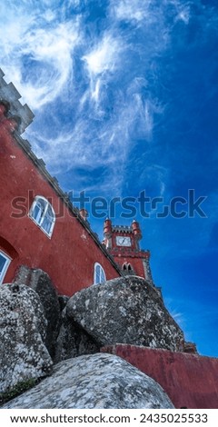 Low angle view of part of a red wall of the Pena Palace on some large rocks under a blue sky with white cobweb clouds backlit by the sun. Sintra. Portugal.