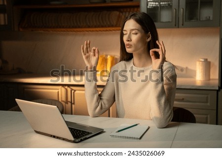 A young woman with her eyes closed set serenely at a wooden table, engaging in a moment of mindfulness before her laptop in the subdued lighting of her home office kitchen. Her fingers are positioned Royalty-Free Stock Photo #2435026069
