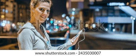 Portrait of smiling young blonde sportswoman listening music on mobile phone application. Banner of female runner with earphones looking at camera after training at night on town. Right copy space.