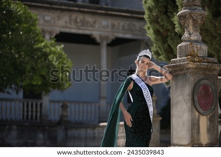 Young, pretty, blonde woman in green party dress with sequins, diamond crown and beauty pageant winner's sash, smiling and happy leaning on a column. Concept of beauty, fashion, pageant, model. Royalty-Free Stock Photo #2435039843
