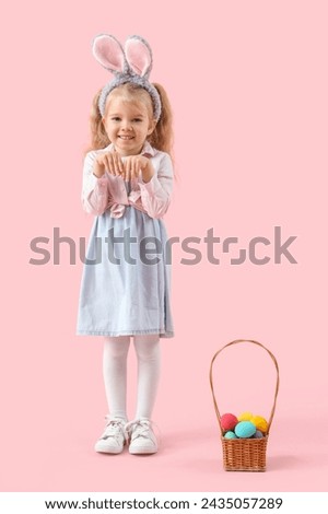 Cute little girl with bunny ears and Easter eggs in wicker basket on pink background