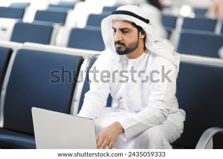 Emirati UAE Dubai man at airport ideal for business trip travel concept. Arab in Kandura dish dasha ghutra. Middle Eastern working using laptop while on the go Royalty-Free Stock Photo #2435059333