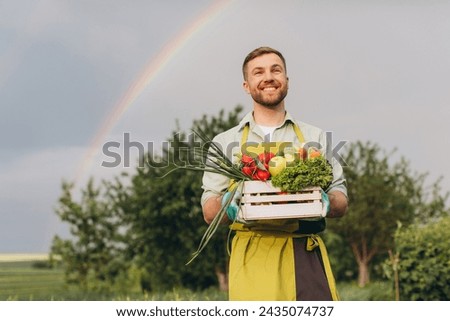 Happy gardener man holding basket with fresh vegetables on rainbow and garden background, gardening concept