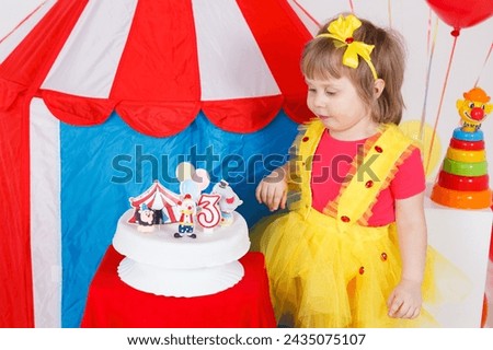A little girl in a colorful outfit is looking at a birthday cake. Circus theme, white, blue, red, yellow colors. Horizontal picture. 