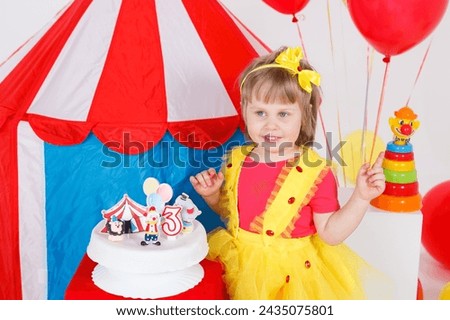 A little girl in a colorful outfit by a birthday cake. Circus theme, white, blue, red, yellow colors. Horizontal picture. 