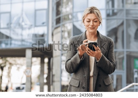 Portrait of a beautiful mature business woman in suit and gray jacket smiling and talking on a phone on the modern urban and office buildings background