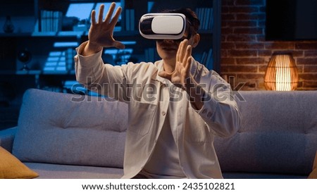 Asian adult man using 3d goggles eyeglasses for playing game at home at night Korean male wear VR glasses helmet play metaverse using virtual reality for business working project at evening at couch