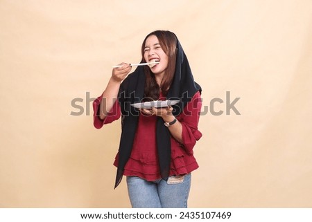 Beautiful mature indonesia woman in red shirt wearing a hijab bites dimsum with chopsticks cheerfully and carries a plate containing dimsum (Chinese food) candid. used for food and culinary content Royalty-Free Stock Photo #2435107469