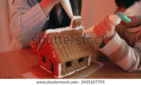Children Adding Royal Icing on Top of Gingerbread House, Family Engaged in Preparing for Holiday Festivities